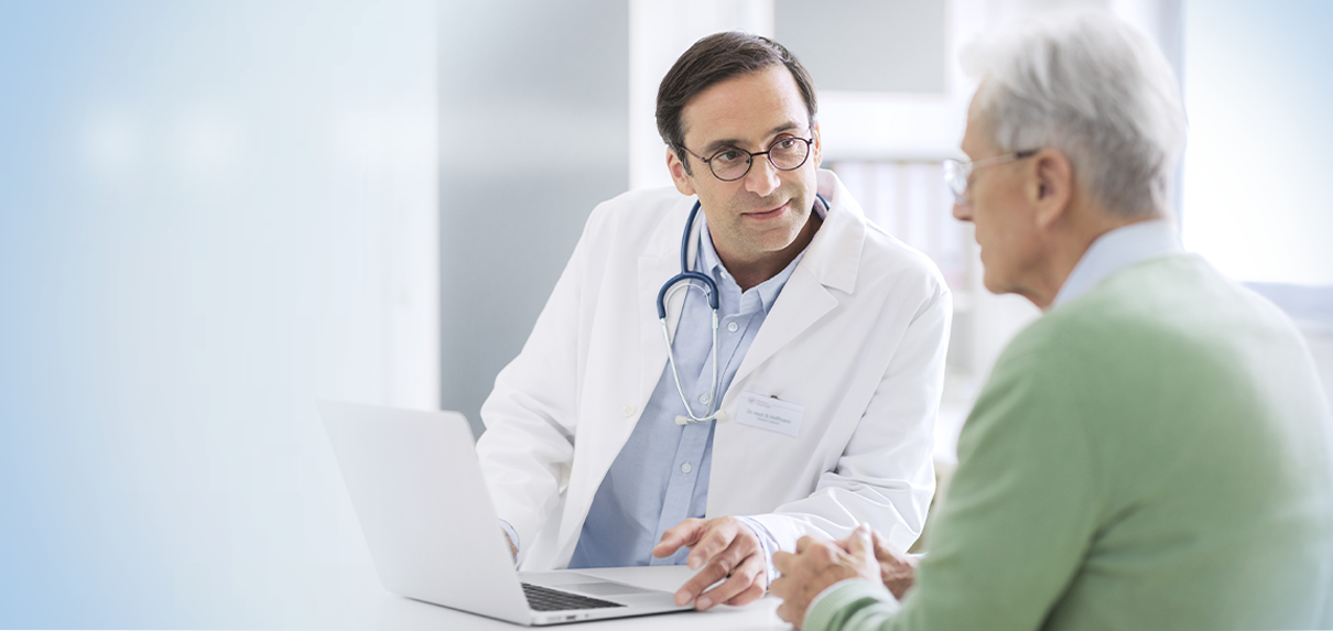 Arzt spricht mit älterem Patienten am Laptop im Sprechzimmer.