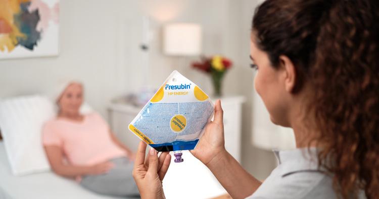 Eine junge Pflegerin zeigt einer älteren Patientin, die zu Hause in ihrem Bett liegt, die Fresubin Sondennahrung, die sie versorgen soll.