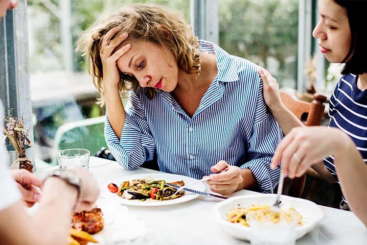 Eine Frau rührt ihr Essen aufgrund von Appetitlosigkeit nicht an, während ihr eine Freundin unterstützend die Hand auf die Schulter legt.