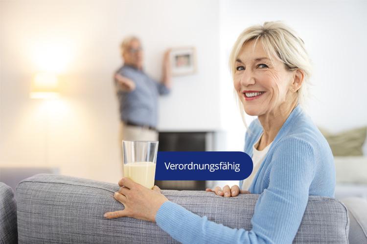 Eine lächelnde Frau sitzt auf einem Sofa und hält ein Glas mit Fresubin Trinknahrung. Ein blauer Button mit der Aufschrift 'Verordnungsfähig' ist eingeblendet.