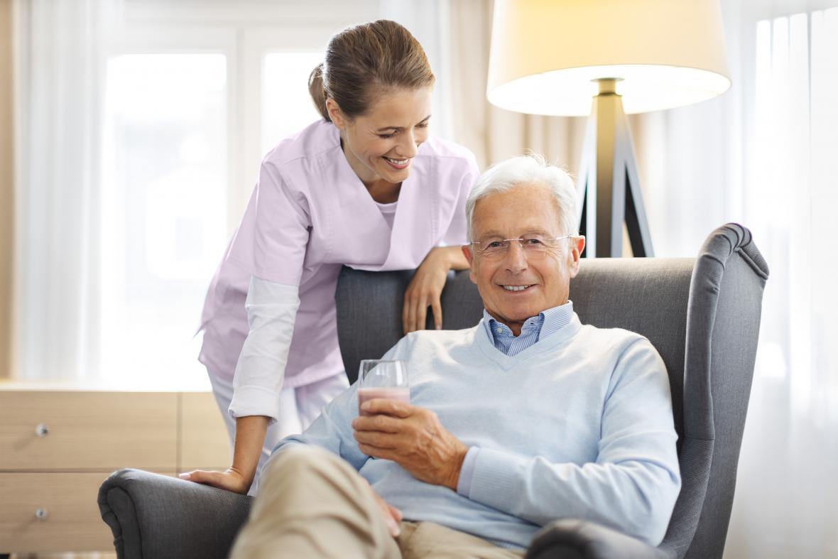 Patient und Pflegerin im Wohnzimmer. Der Patient hält ein Glas mit Fresubin Trinknahrung in der Hand.