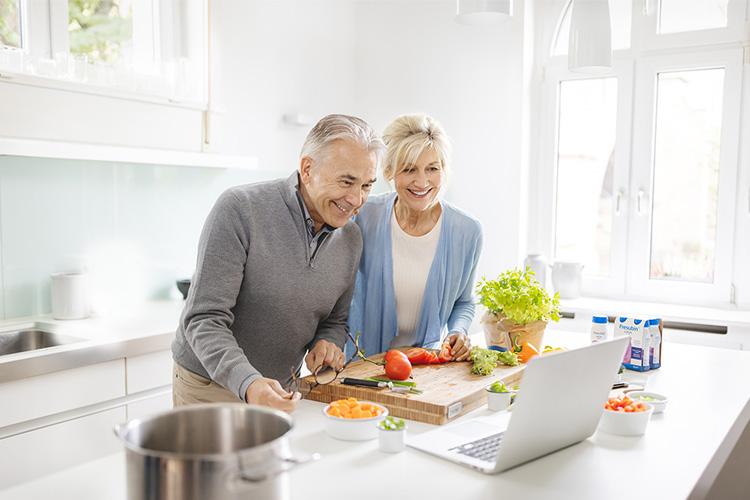 Mangelernährung im Alter: Älteres Paar schaut auf Laptop und kocht gemeinsam mit Gemüse und Fresubin Trinknahrung -verlinkt zu Rezepte