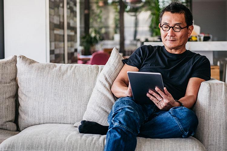 asian senior on couch with his tablet reading 